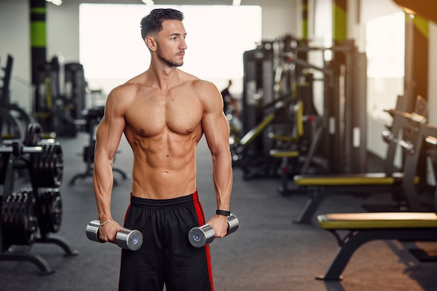 Hombre fitness con torso desnudo trabajando con pesas durante el entrenamiento de manos y hombros en el gimnasio. Concepto deportivo y saludable.