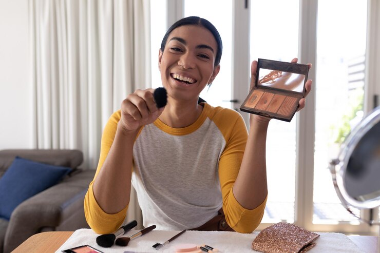 Foto hombre fluido de género de raza mixta haciendo vlog sosteniendo maquillaje en polvo y pincel, sonriendo