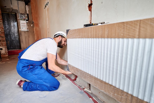 Hombre fontanero en mono de trabajo instalando un radiador de calefacción en una habitación vacía