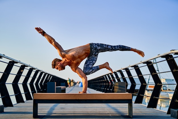 Hombre en forma haciendo un soporte con una mano mientras practica yoga solo cerca del océano contra el cielo al atardecer o al amanecer