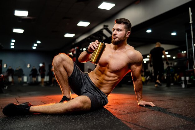 Hombre en forma de músculo en el gimnasio relajado bebiendo cócteles de proteínas. Chico sentado en el suelo en el gimnasio moderno.