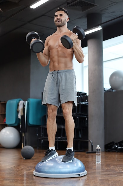 Hombre en forma y musculoso trabajando con bumbbells en bola bosu de hemisferio gimnástico en el gimnasio.