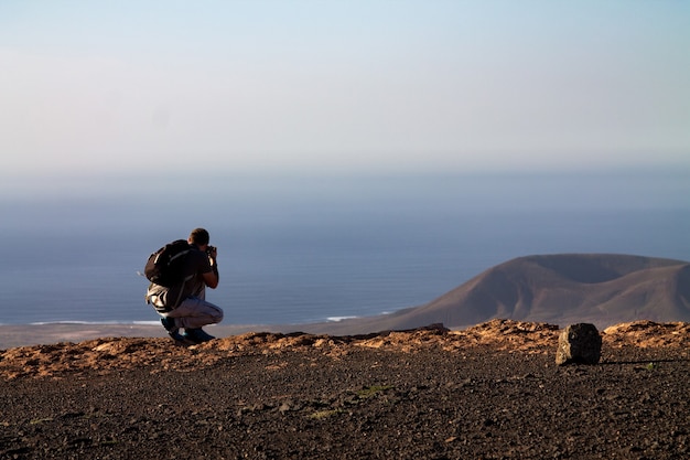 Un hombre fotografía una isla en el océano, en cuclillas al borde de un acantilado. Mirador del Rio, Lanzarote, España.