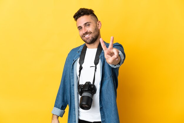 Hombre fotógrafo aislado en amarillo sonriendo y mostrando el signo de la victoria