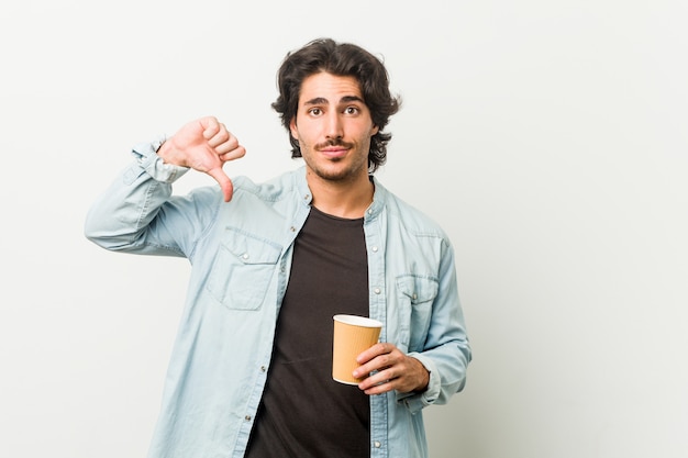 Hombre fresco joven que bebe un café que muestra un gesto de la aversión, pulgares abajo. Concepto de desacuerdo