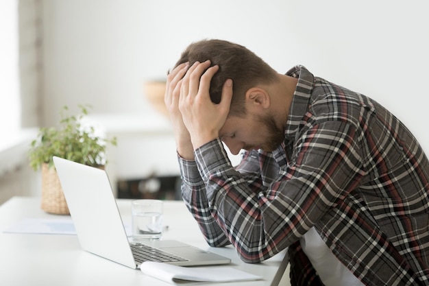 Hombre frustrado deprimido con la cabeza en las manos conmocionado por la bancarrota caída de las acciones sentado en el escritorio de trabajo con la computadora portátil estresado cansado hombre de negocios se siente desesperado dinero perdido en línea o tiene problemas de deuda