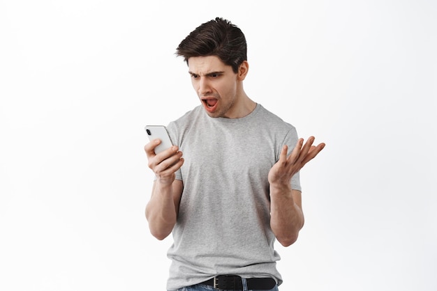 El hombre frustrado se ve confundido en la pantalla del teléfono inteligente, lee un mensaje extraño en el teléfono móvil y argumenta que se queja de pie contra el fondo blanco
