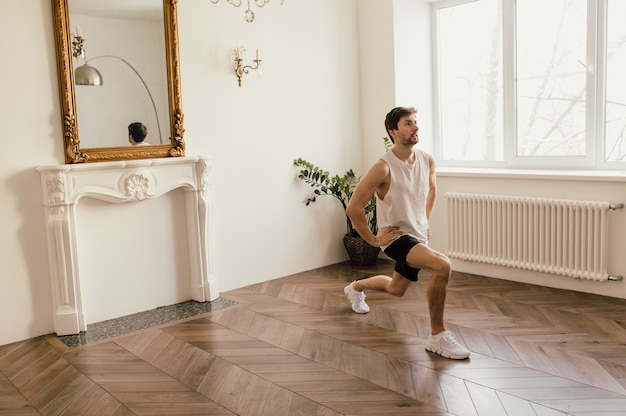 Hombre fuerte atlético en camiseta y pantalones cortos está haciendo ejercicios de sentadillas en casa en su espacioso y br ...