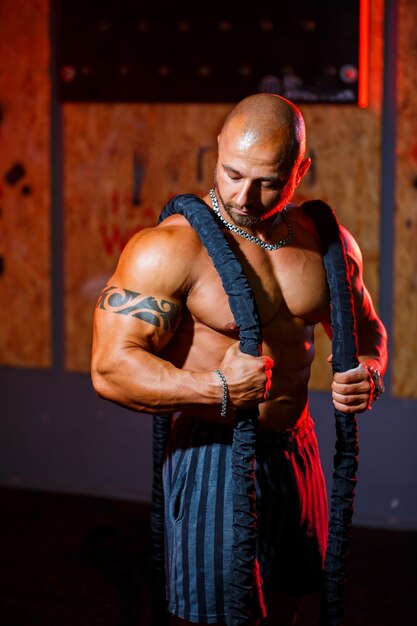 Hombre fuerte guapo deportivo posando con cuerda de deportes en el fondo del gimnasio. Un culturista fuerte con abdominales, hombros, bíceps, tríceps y pecho perfectos.