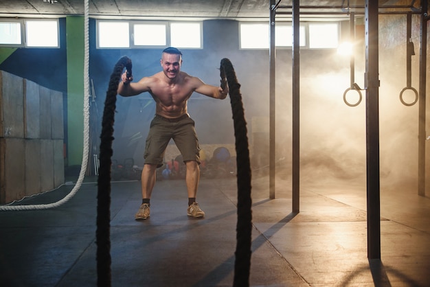 Hombre fuerte guapo haciendo ejercicios para los músculos con cuerdas de batalla en el gimnasio.