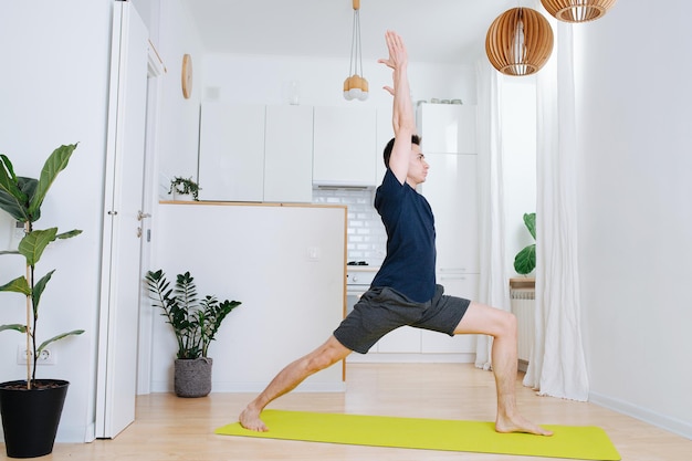 Hombre fuerte y robusto haciendo guerrero asana practicando yoga en la cocina de casa