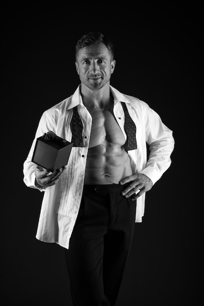 Hombre fuerte con torso abdominal de seis paquetes en camisa abierta sostenga el regalo de fondo negro de la caja actual