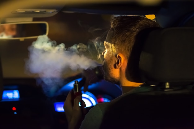 El hombre fuma un cigarrillo electrónico en el auto. Por la noche, por la noche