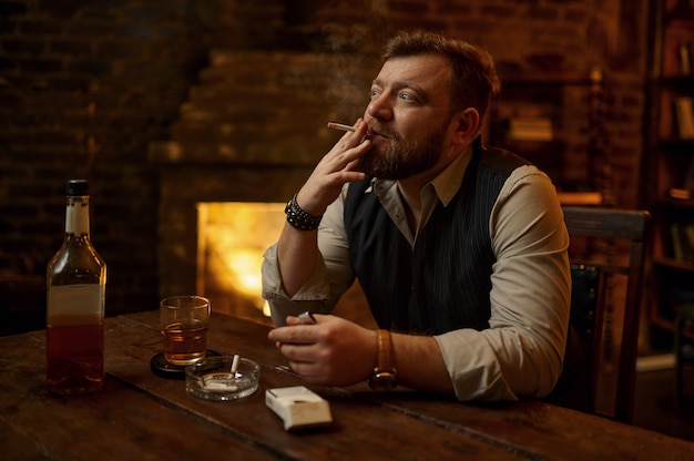 El hombre fuma cigarrillos y bebe bebidas alcohólicas, estantería y rico interior de oficina. Cultura del tabaquismo, sabor específico