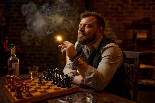 El hombre fuma un cigarro, bebe bebidas alcohólicas y juega al ajedrez, estantería y el interior de la oficina vintage. Cultura del tabaquismo, sabor específico. Ocios de fumador masculino en el tablero de ajedrez