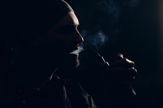 Hombre fumando una pipa en la oscuridad. Retrato de perfil retroiluminado.