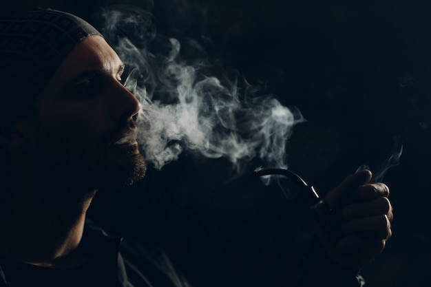 Hombre fumando una pipa sobre fondo oscuro. Retrato de perfil retroiluminado.