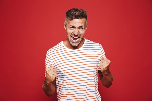 Foto hombre furioso emocional en camiseta a rayas gritando y mostrando puños aislados en rojo