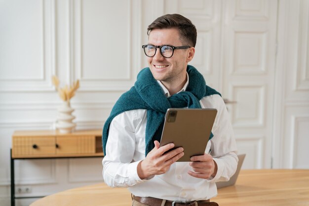 Un hombre con gafas y una bufanda azul azul se para sosteniendo una tableta mirando lejos pensativo