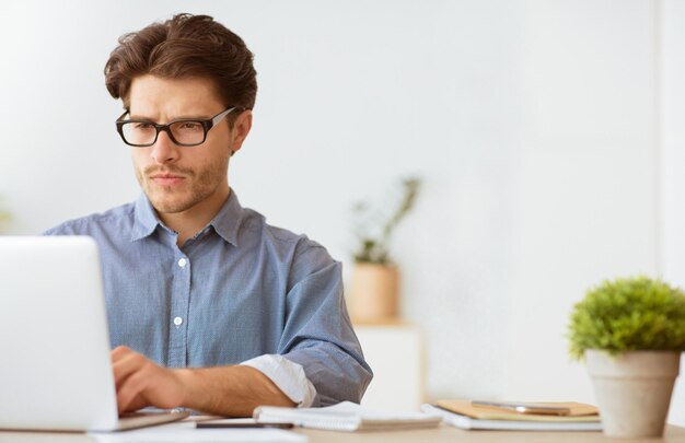 Un hombre con gafas está sentado en un escritorio en una oficina centrado en su computadora portátil