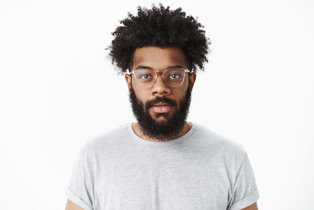 hombre de gafas con nariz perforada y peinado afro sonriendo alegremente sintiéndose positivo, de pie contra el gris.