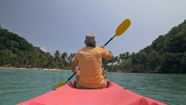 Hombre con gafas de sol filas de canoa de plástico rosa a lo largo del mar contra verdes islas montañosas con selvas salvajes