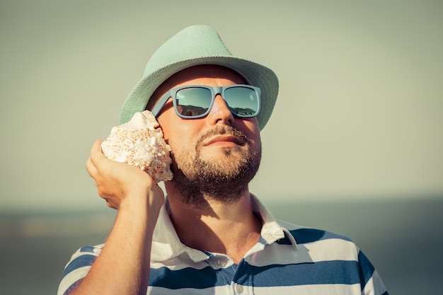 Hombre con gafas de sol y un sombrero escuchando una concha escuchar concha contra el mar y el fondo del cielo