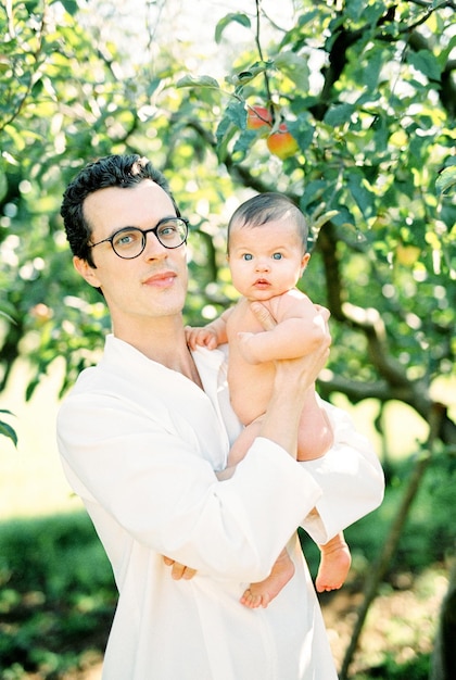 Hombre con gafas sostiene a un bebé en sus brazos mientras está parado debajo de un manzano en el jardín