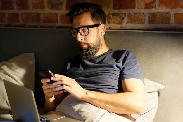 Hombre con gafas usando teléfono y portátil en la cama