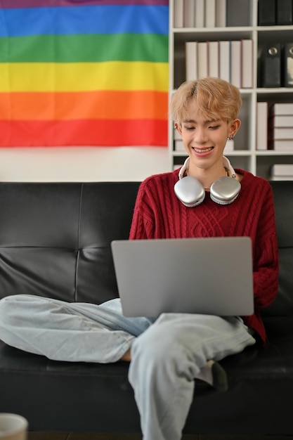 Un hombre gay asiático feliz está usando su computadora portátil mientras se relaja en un sofá en su sala de estar