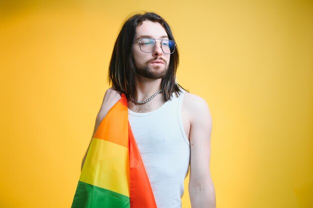 Hombre gay con una bandera del orgullo gay sonriendo y mirando hacia otro lado sobre fondo amarillo