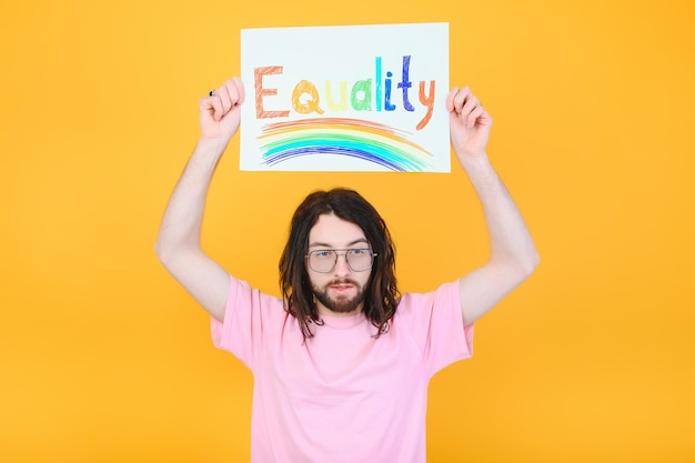 Hombre gay con una bandera del orgullo gay sonriendo y mirando hacia otro lado sobre fondo amarillo