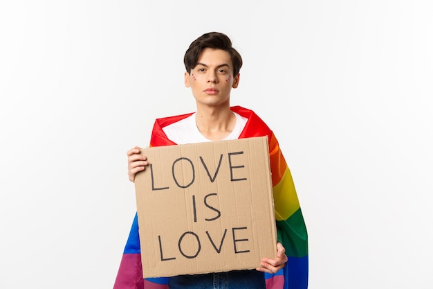 Foto hombre gay serio y seguro con la bandera lgbt del arco iris, con cartel para el desfile del orgullo, de pie sobre blanco.