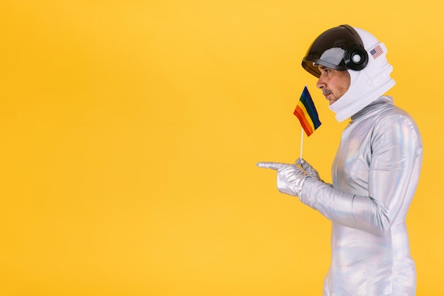 Hombre gay vestido de astronauta con casco y traje plateado con una bandera del colectivo lgtbi apuntando con las manos sobre un fondo amarillo Concepto gay homosexual y trans