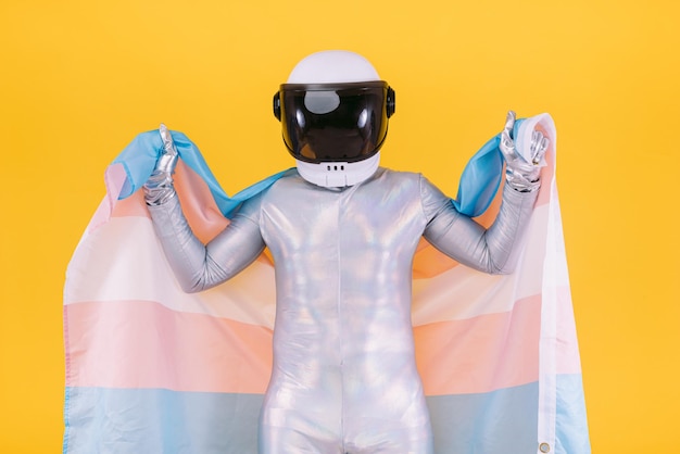 Hombre gay vestido como astronauta con casco y traje plateado sosteniendo una bandera de la comunidad trans sobre un fondo amarillo Derechos homosexuales homosexuales trans y concepto de orgullo de género