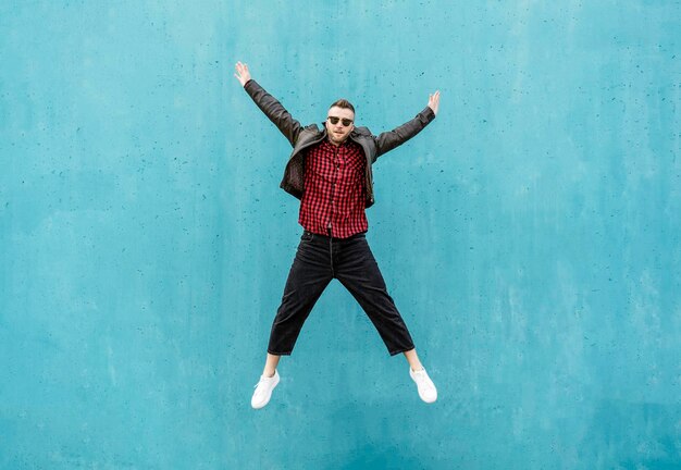 Un hombre genial con ropa indie casual saltando al aire libre con los brazos abiertos contra un fondo de pared azul Un chico de moda divirtiéndose bailando y celebrando al aire libre Concepto de estilo de vida para hombres maduros alegres