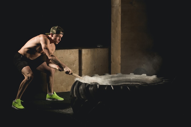 El hombre golpea a Tyre. Entrenamiento en el gimnasio con martillo y neumático de tractor