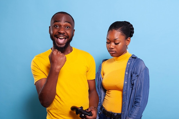 Hombre golpeando a mujer en videojuegos con controlador en consola sobre fondo azul. El novio celebra la victoria y la novia se siente decepcionada por perder el juego en línea en el estudio.