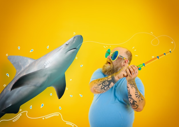 Foto hombre gordo asustado con barba y gafas de sol atrapó a un tiburón con la caña de pescar