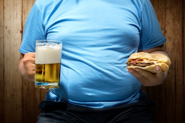 Hombre gordo con cervezas y hamburguesa en la mano.