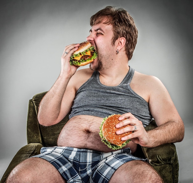 Hombre gordo comiendo hamburguesa sentado en un sillón. Estilo de comida rápida.