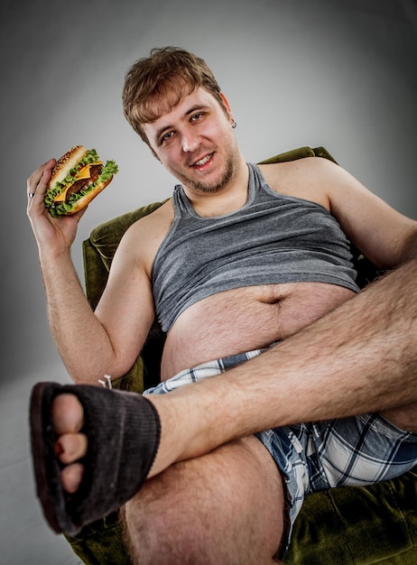 Hombre gordo comiendo hamburguesa sentado en un sillón. Estilo de comida rápida.