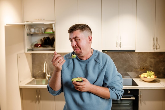 Hombre gordo y divertido comiendo aguacate mientras está de pie en la cocina casera moderna Concepto de estilo de vida saludable y adelgazante