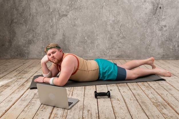 hombre gordo divertido haciendo deporte en la estera de yoga, haciendo deportes en línea, mirando a la computadora portátil.