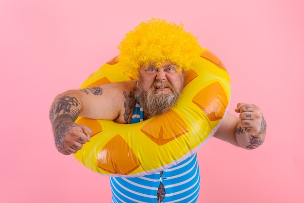 Hombre gordo enojado con peluca en la cabeza está listo para nadar con un salvavidas de rosquilla