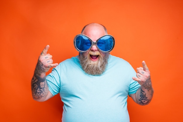 Hombre gordo feliz con tatuajes de barba y gafas de sol hace el gesto de los cuernos