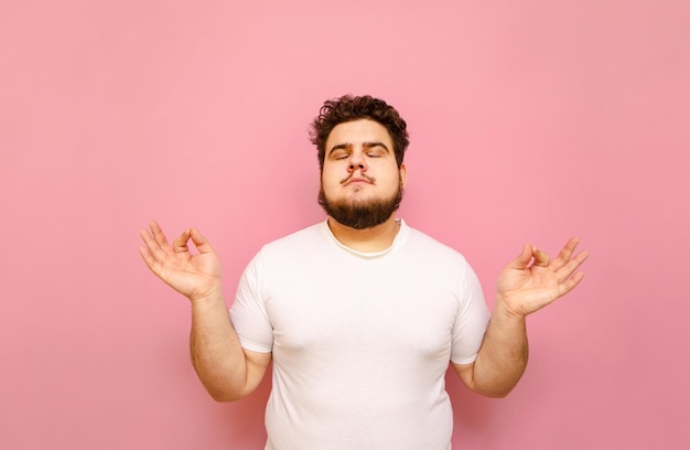 Hombre gordo guapo con pelo rizado y barba medita sobre un fondo rosa con los ojos cerrados