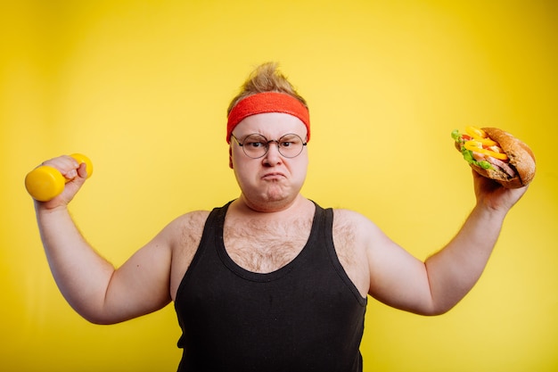 Hombre gordo hambriento mostrando bíceps con hamburguesa y pesa
