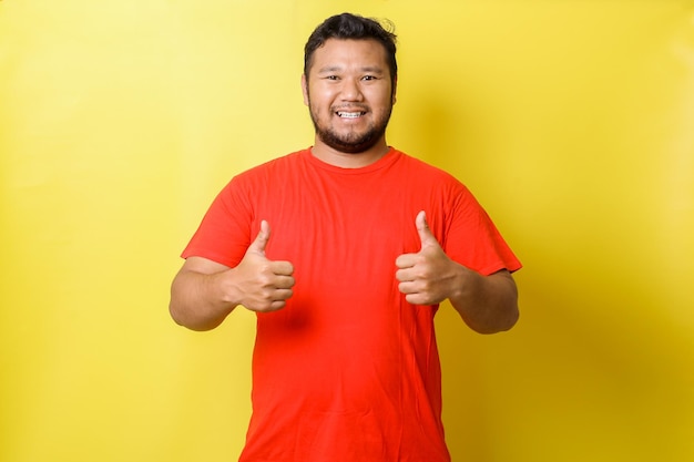 Hombre gordo joven asiático que muestra dos anuncios publicitarios de pulgar doble solución de confianza aislada sobre centrico amarillo