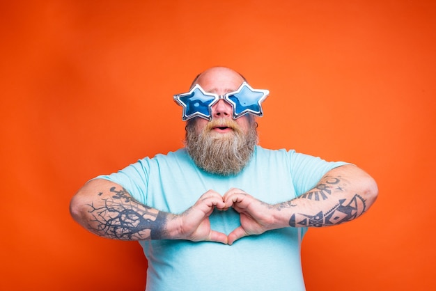 Hombre gordo con tatuajes de barba y gafas de sol hace con las manos en forma de corazón